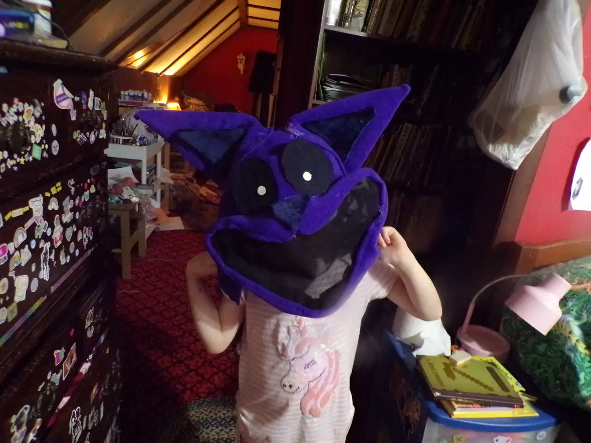 A person in a child's bedroom, wearing a large purple bat costume mask with big black eyes and pointed ears, holds the mask…