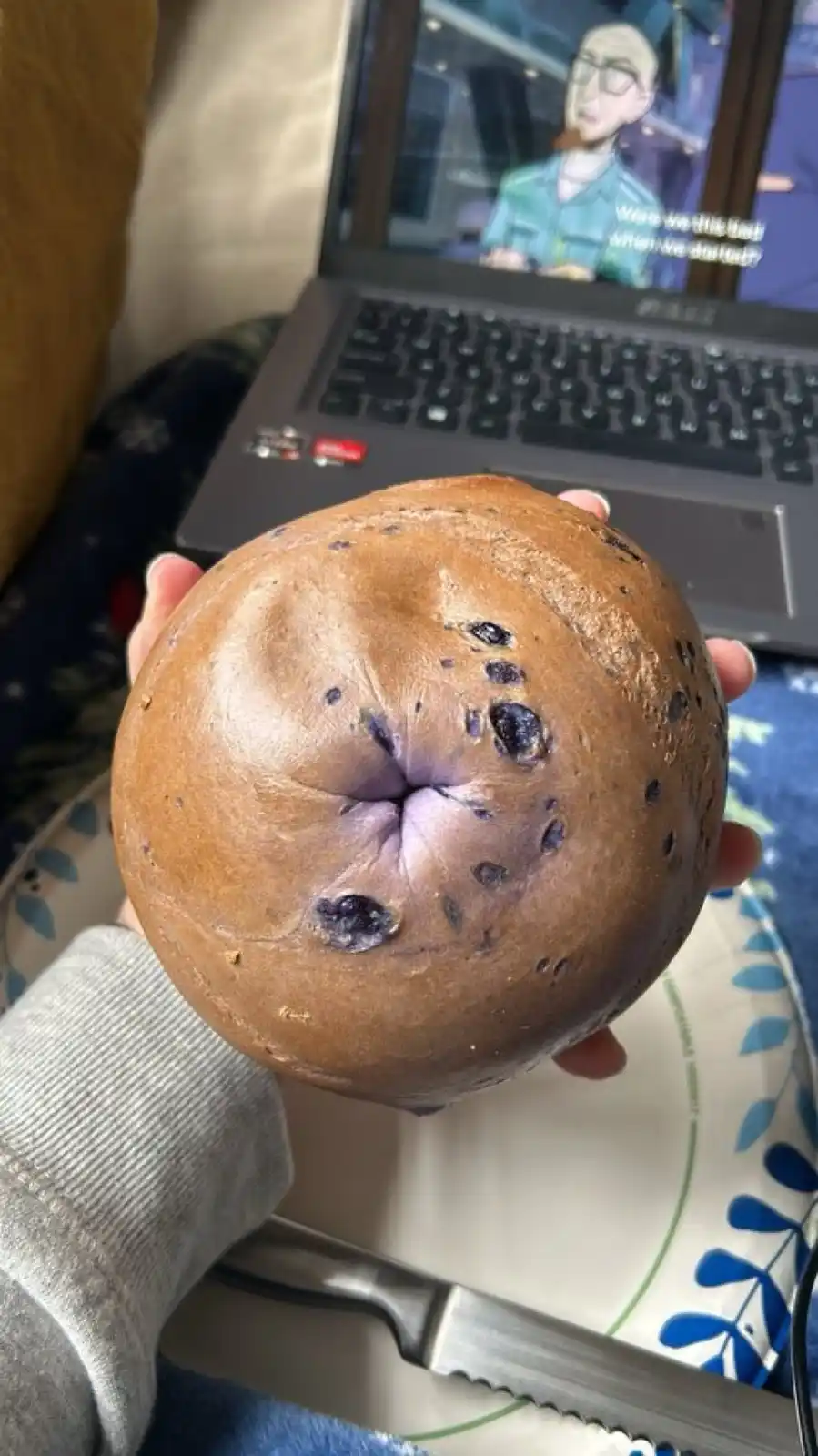 A hand holds a round, brown bagel with a purple center and blueberry-like spots, resembling a human face. In the background…