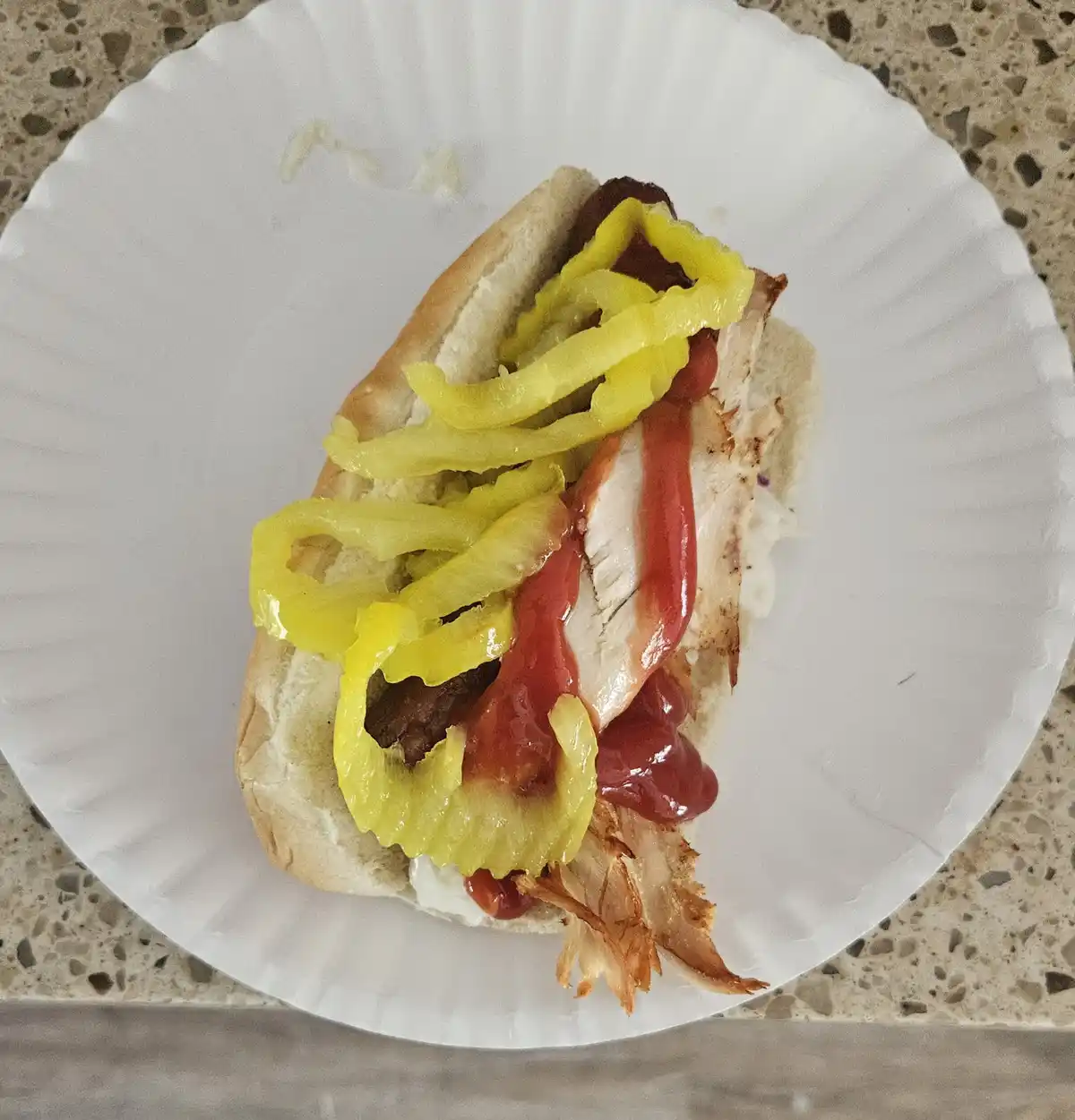 A hot dog bun sits on a white paper plate, filled with shredded chicken, ketchup, and yellow pickled peppers, with a small…