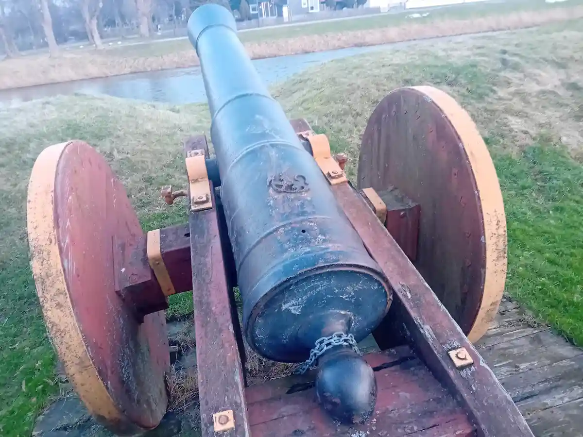 A weathered, blue-painted historical cannon with large wooden wheels sits on grass near a canal, its cannonball chained to…