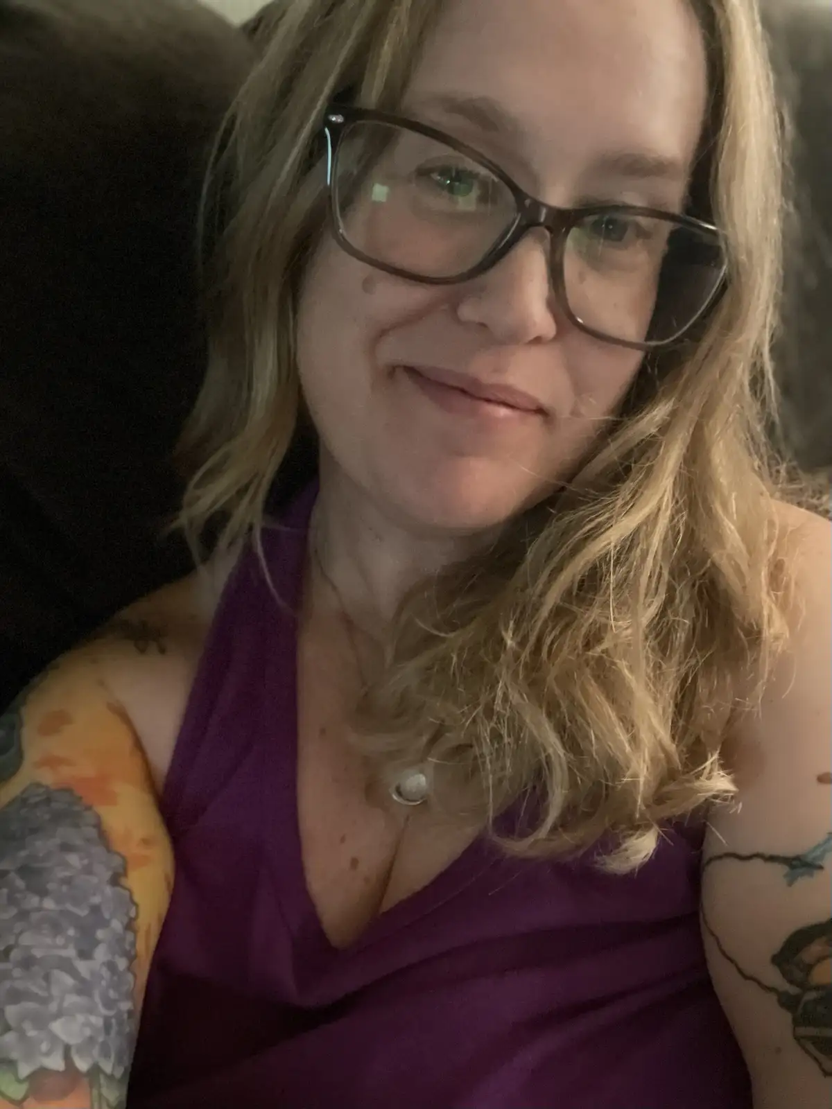 A woman with blonde hair and glasses smiles slightly while wearing a purple top, showing tattoos on both arms. The overlaid…