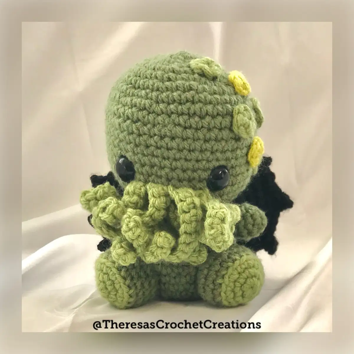A small, green, crocheted plush toy resembling the fictional creature Cthulhu, featuring large black eyes, black wings, and…
