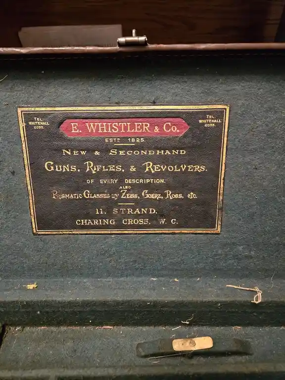 A close-up of a vintage, dark green gun case interior showing a brass plaque for 'E. WHISTLER & Co.', established in 1825…