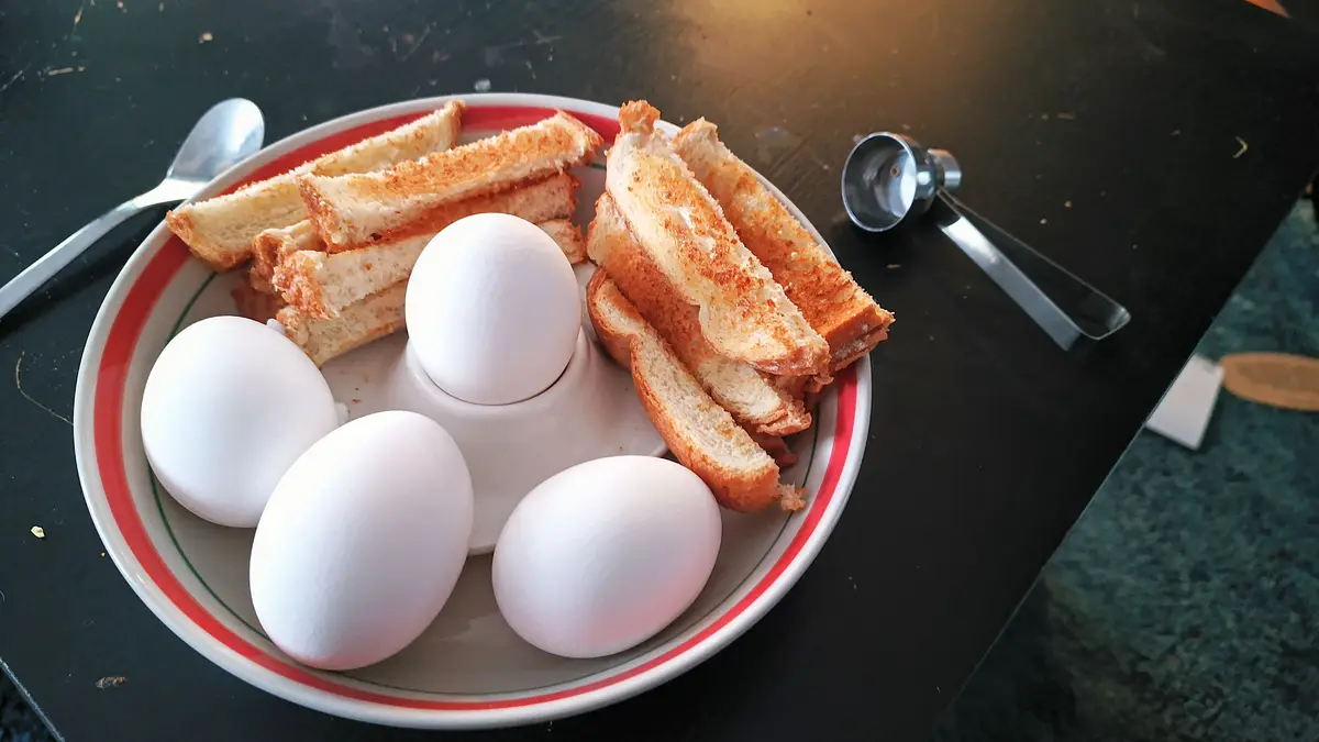A plate of breakfast food including four whole white eggs and several slices of toasted bread, with a metal egg cup holding…