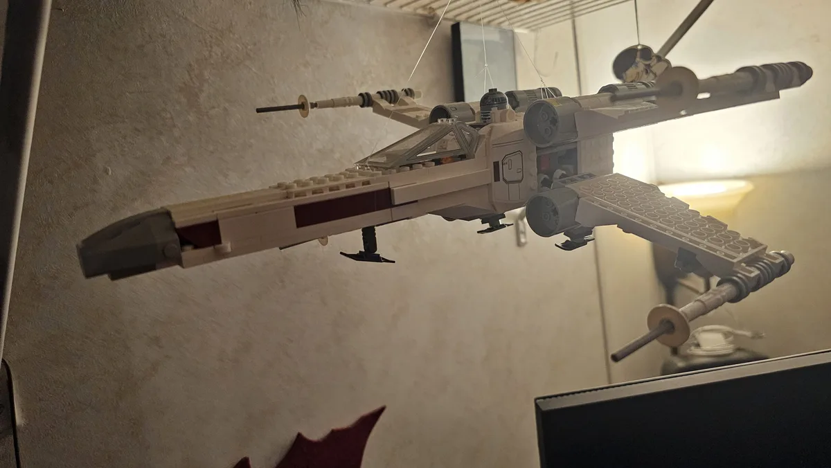 A LEGO Star Wars X-wing fighter model is suspended in mid-air by thin strings, appearing to float in a room with a textured…