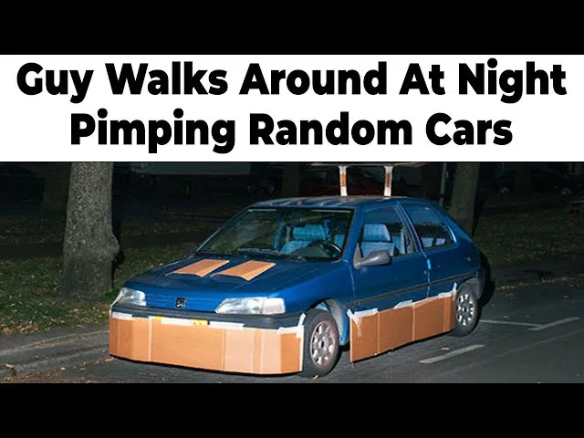 A blue car is parked at night with cardboard boxes taped to its front bumper and side panels, resembling a makeshift…