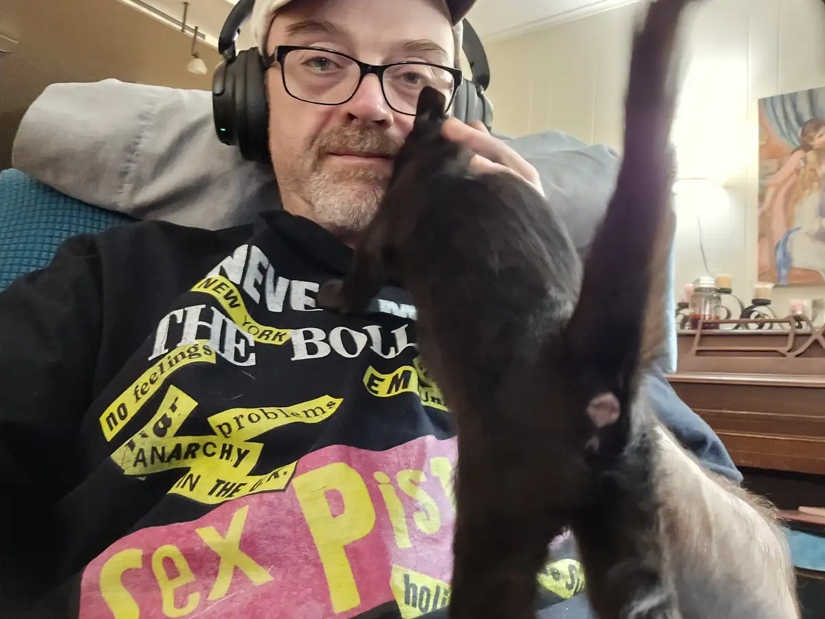 A man wearing glasses and headphones sits in a chair, holding a black cat that is stretching upward with its front paws…