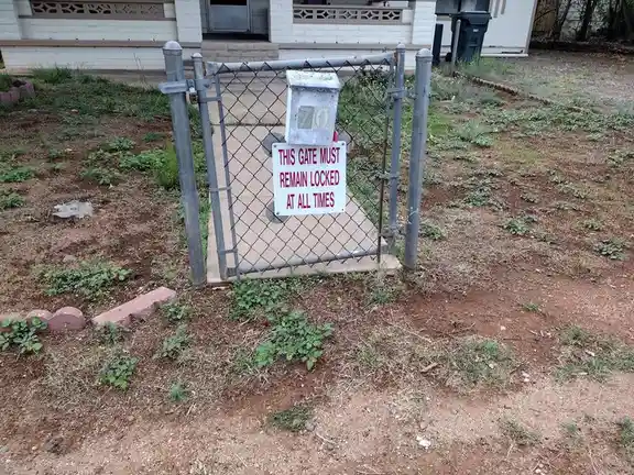 A small chain-link gate stands in a dirt yard with sparse weeds, enclosing a concrete pad where a white utility box is…