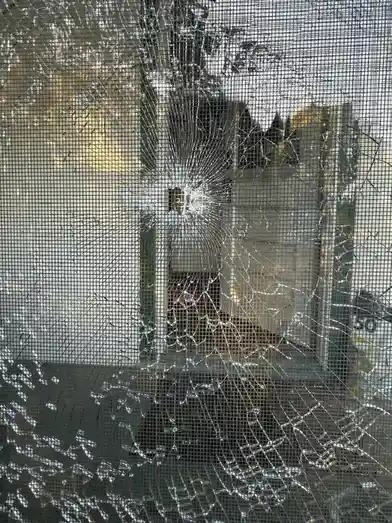 A close-up photo shows a shattered glass window with a screen door mesh overlay, cracks radiating from a central impact…