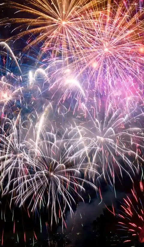 A vibrant nighttime photograph of colorful fireworks exploding in the sky, featuring bursts of red, white, blue, and gold…
