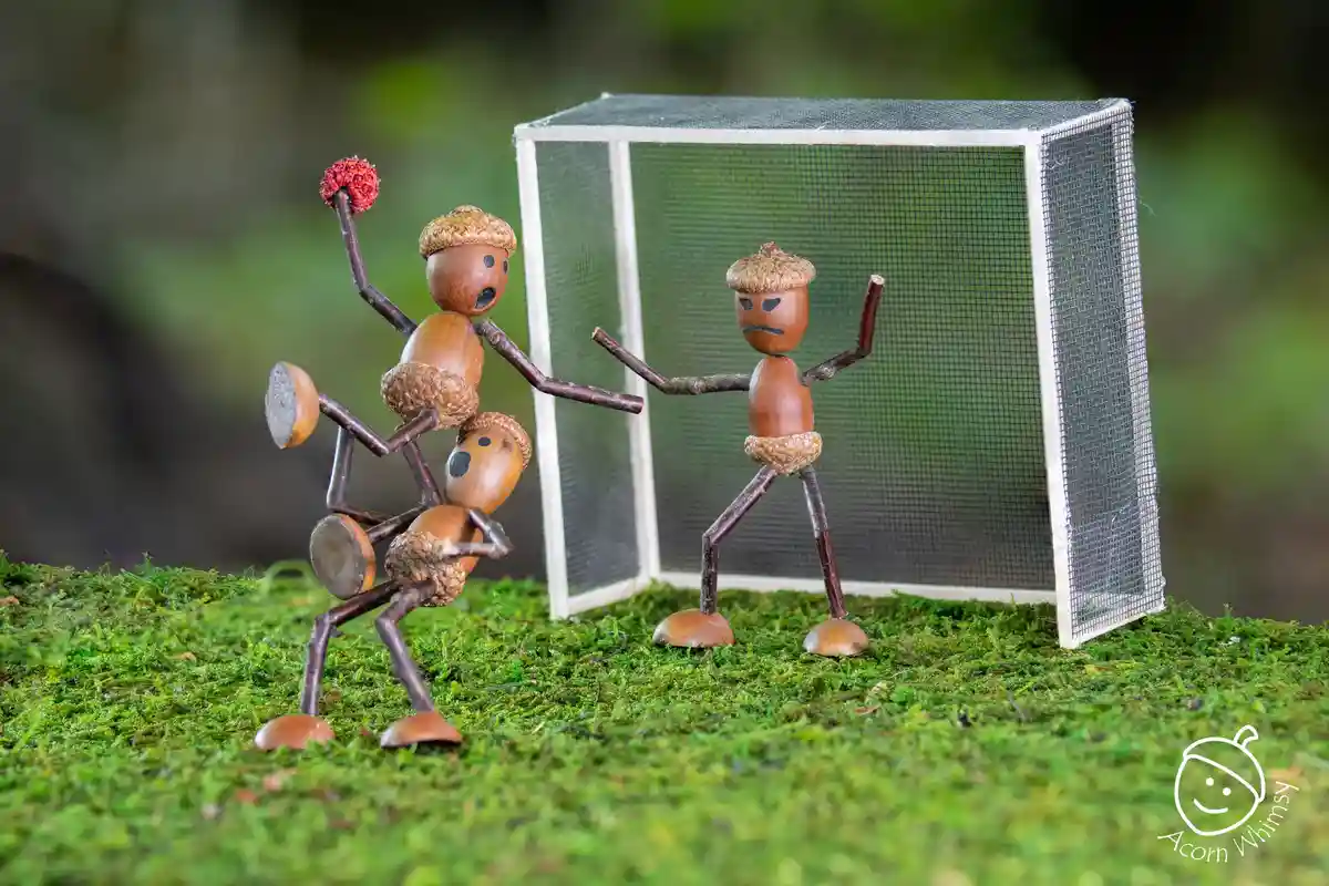 Two handmade acorn figures with twig limbs are staged on mossy ground in front of a miniature soccer goal. The figure on the…