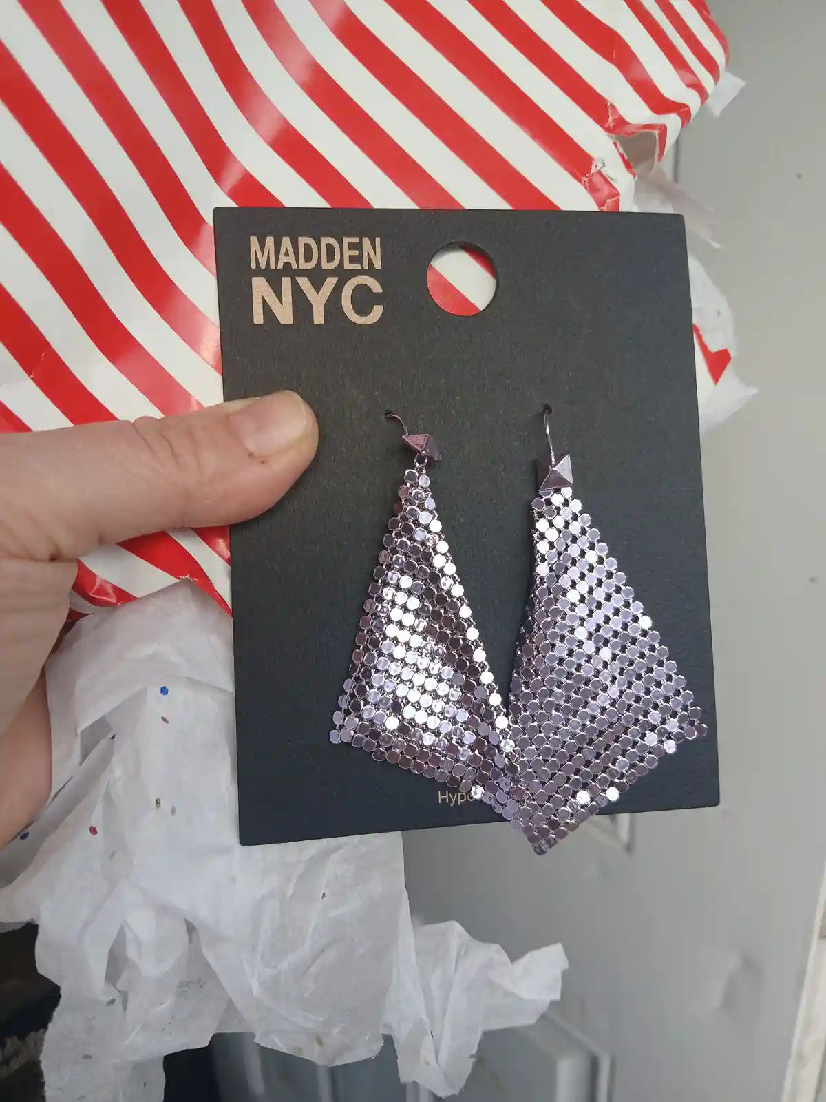 A hand holds a black card displaying a pair of shiny, sequined, tree-shaped earrings from Madden NYC, with red and white…