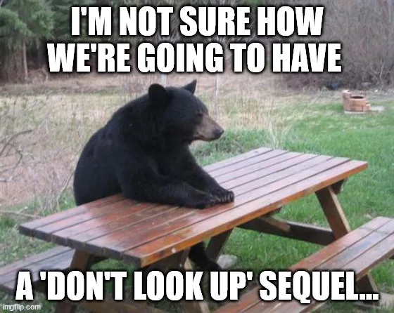 A black bear is sitting on a wooden picnic table outdoors, looking off to the side with a contemplative or concerned…