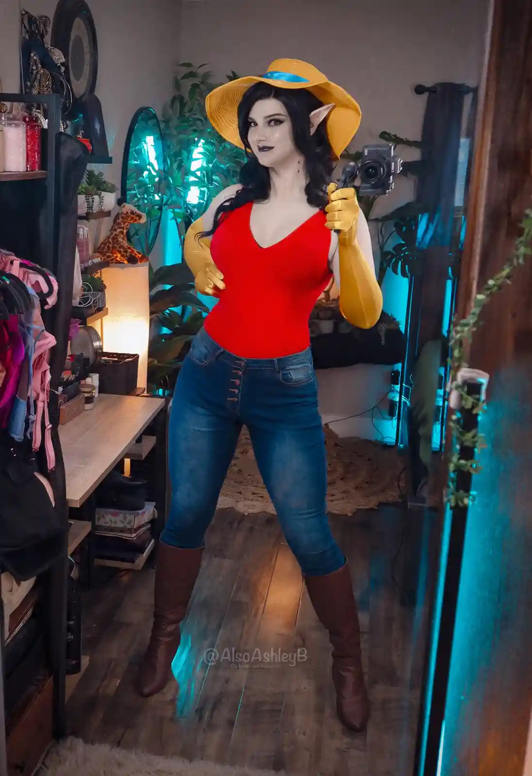 A person with pointed ears and dark makeup poses in a mirror selfie, wearing a yellow sun hat, red top, blue jeans, and…