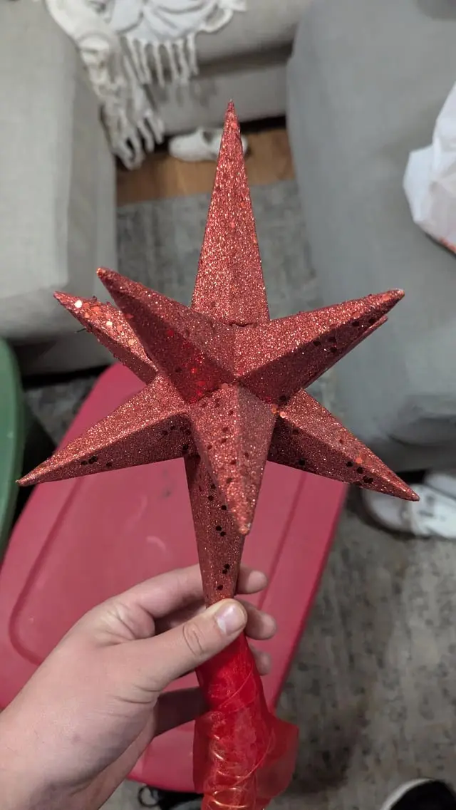 a hand holds a red glittery christmas tree topper shaped like a star, which has multiple small holes punched through its…