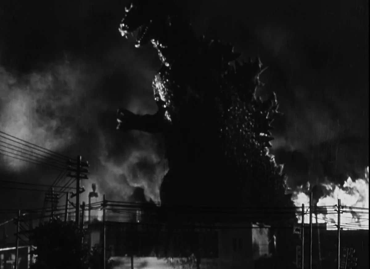 A black-and-white still from the 1954 film 'Godzilla' shows the titular monster towering over a burning city at night…