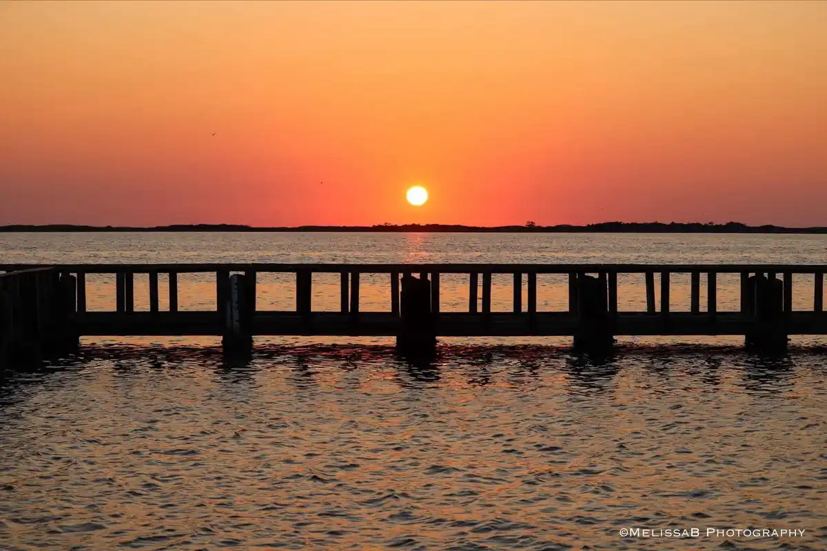 A serene sunset photo shows a wooden pier silhouetted against an orange and red sky with the sun low on the horizon over…