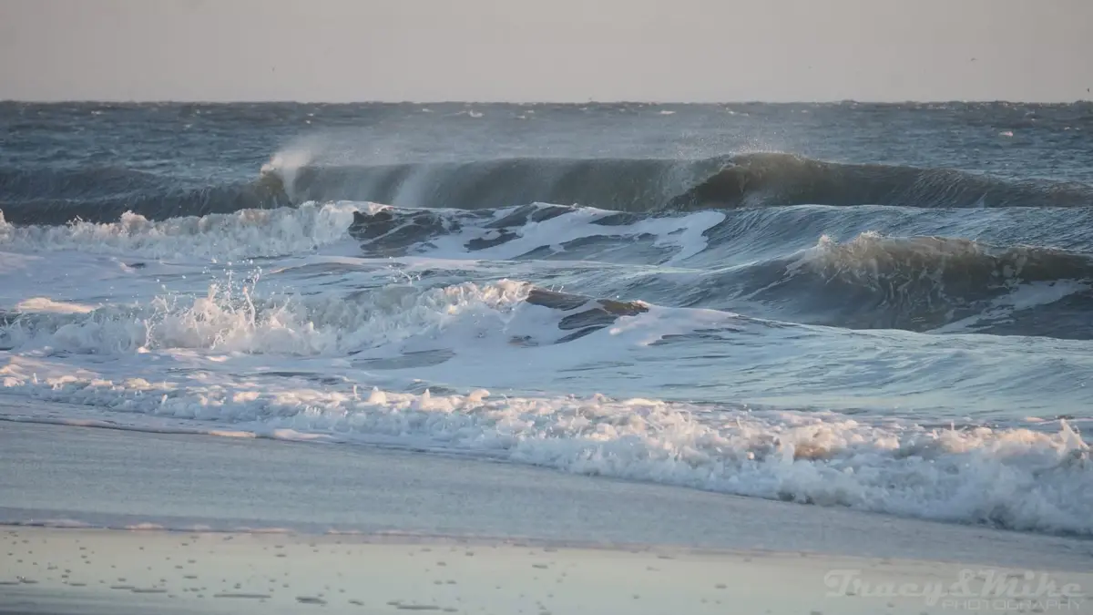 A photograph of turbulent ocean waves crashing onto a beach with white foam, under a hazy sky. The image has a watermark in…