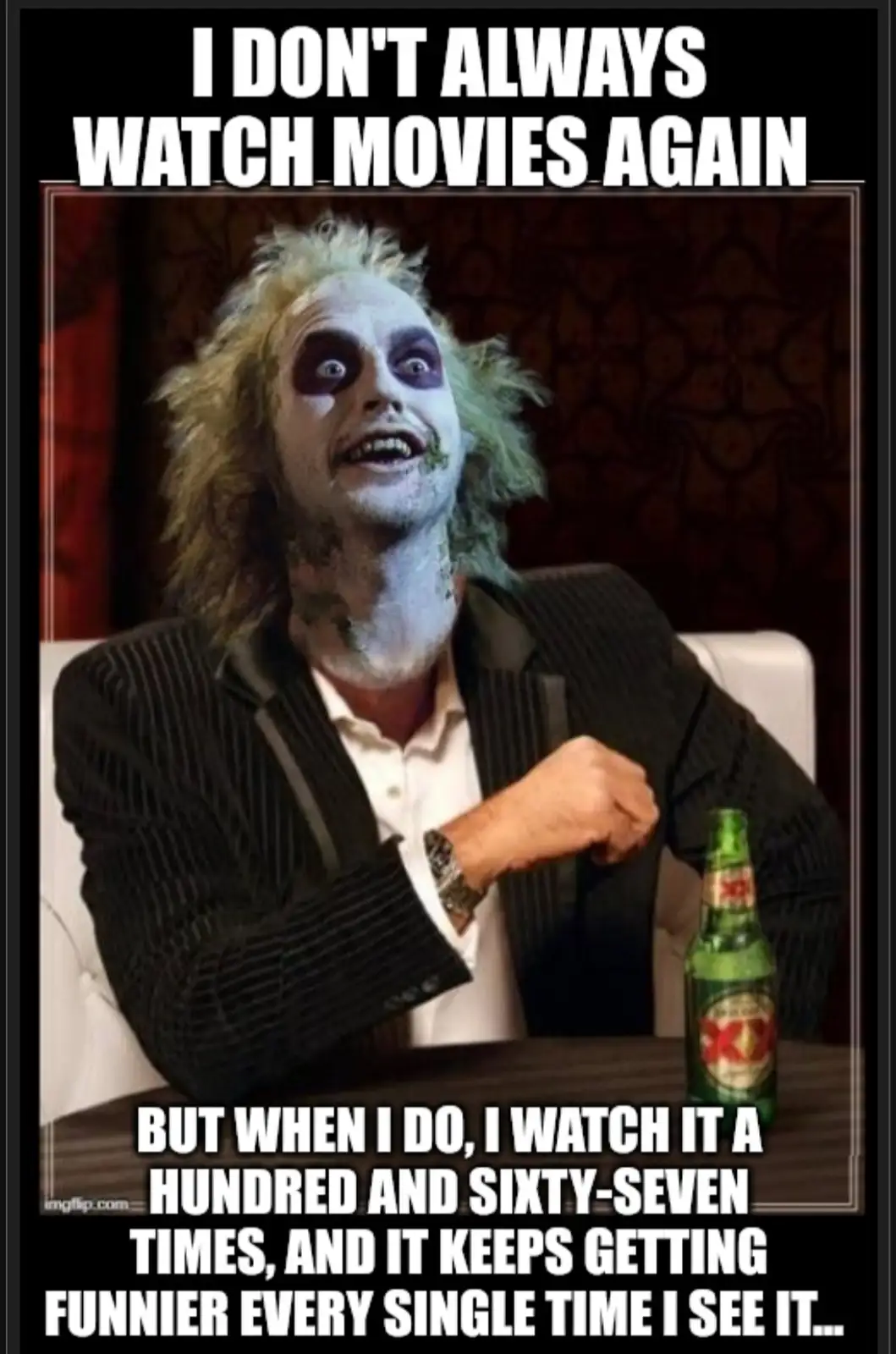 A man with pale makeup, dark eye makeup, and messy hair sits at a table with a green beer bottle. He has a wide, manic grin…