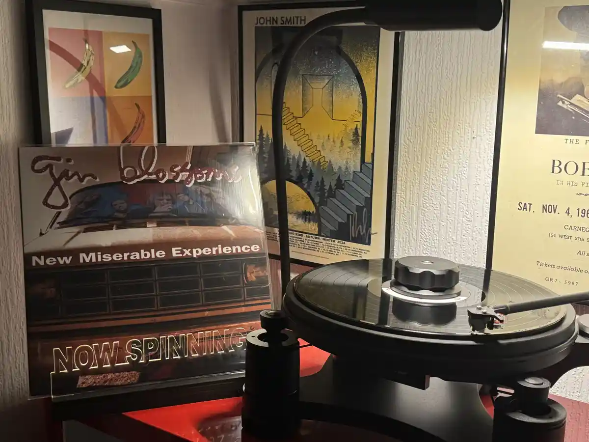 A vinyl record spins on a turntable, with a clear acrylic sign in front of it that reads 'NOW SPINNING'. Behind the…