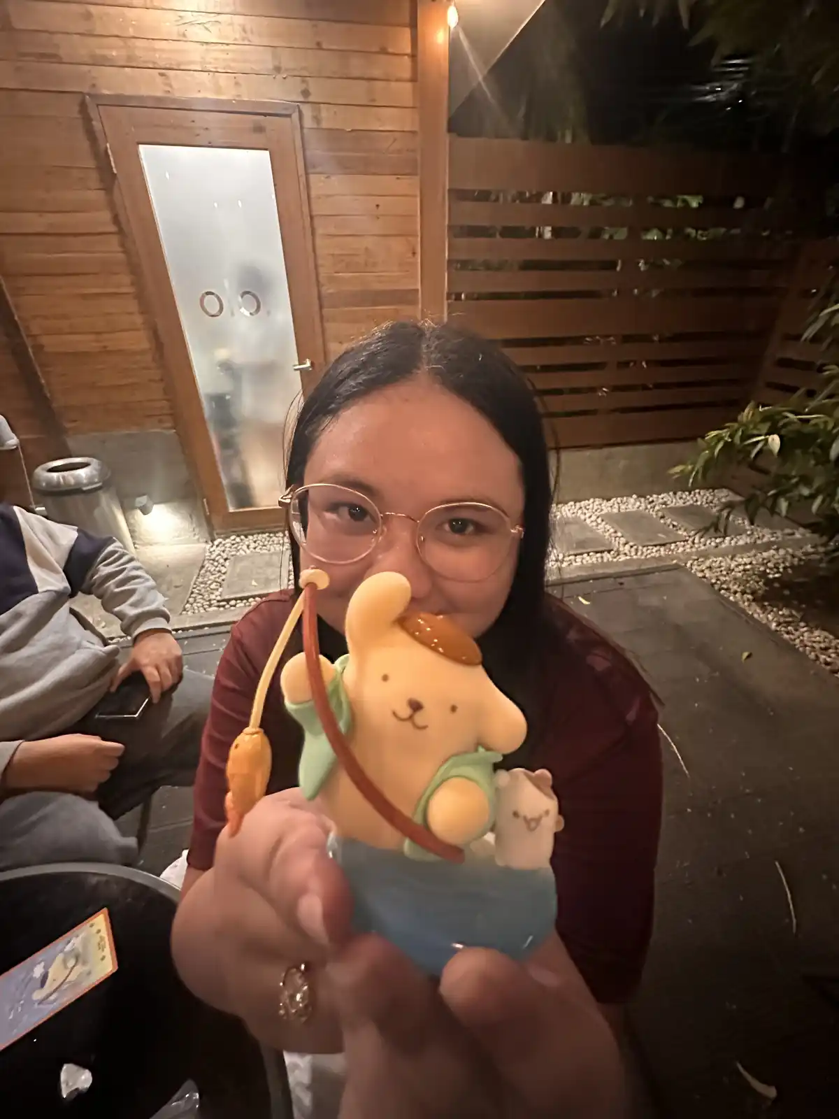 A person with glasses holds up a small figurine of a yellow bunny holding a fishing rod with a carrot as bait, smiling at…