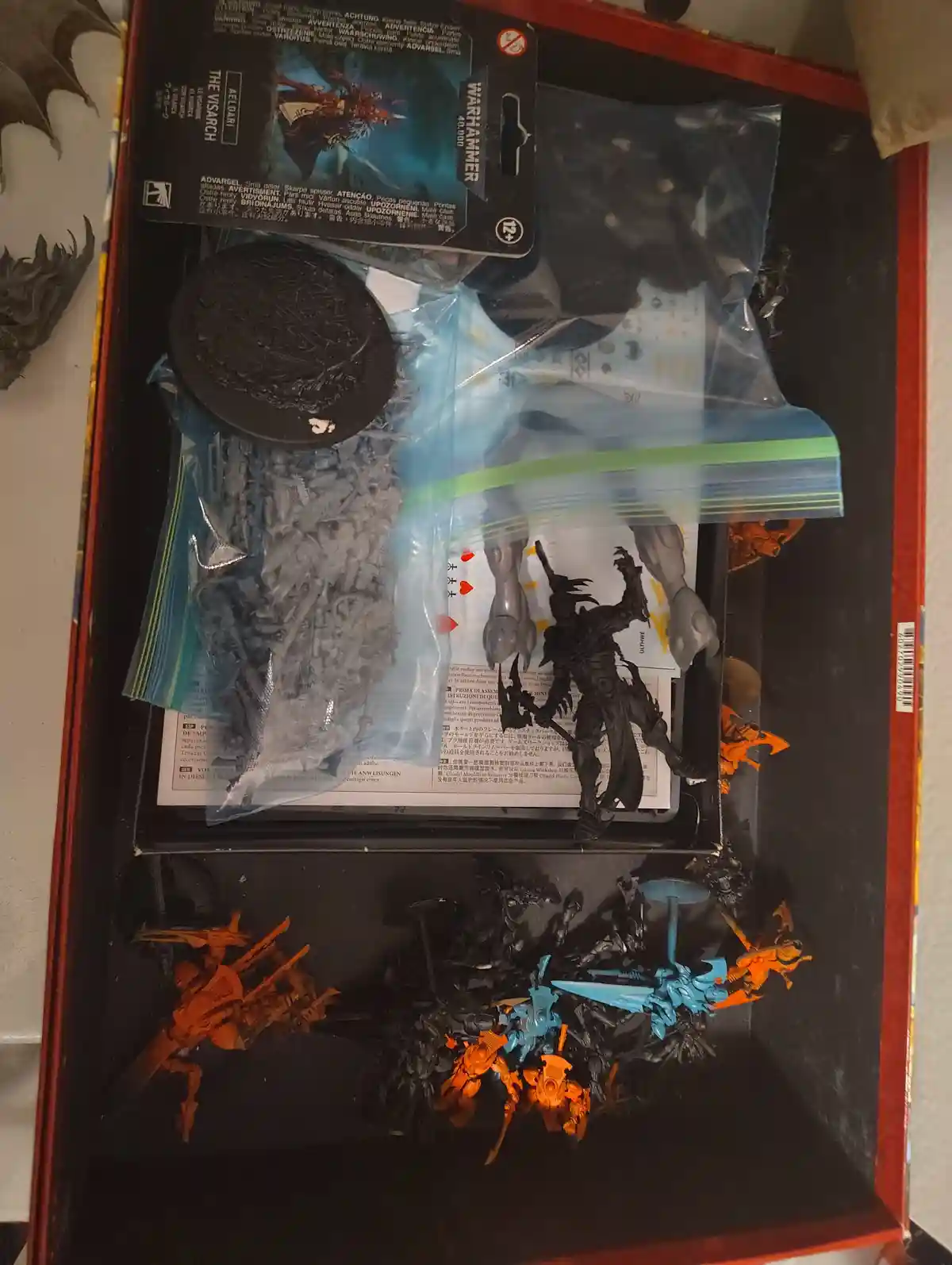 An overhead shot of a Warhammer 40,000 miniatures box containing unassembled grey plastic parts in a ziplock bag, a painted…