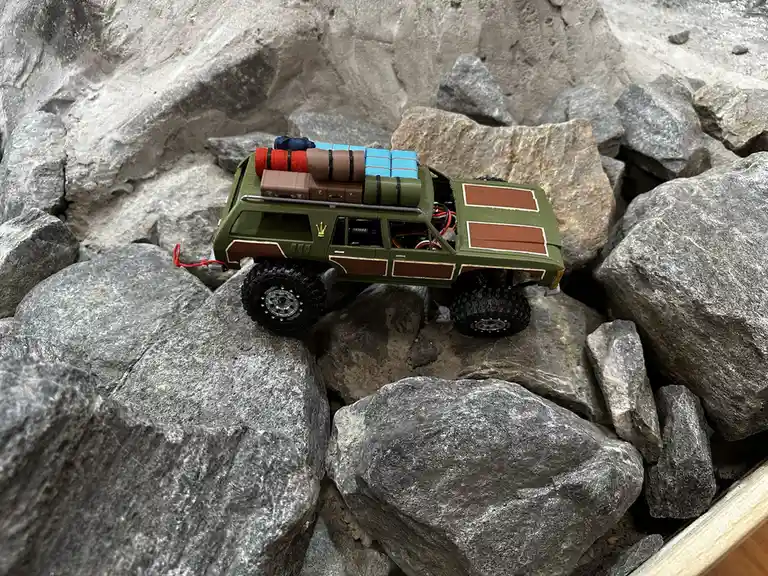A green and brown toy SUV with large off-road tires and cargo strapped to its roof is stuck among large gray rocks. The…