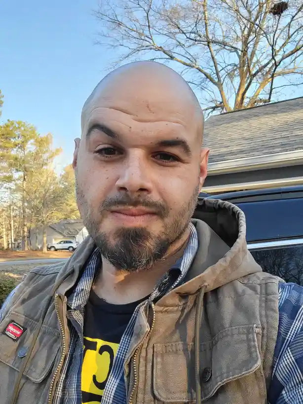 A bald man with a goatee and a slight smirk takes a selfie outdoors in front of a house and a dark SUV. He wears a plaid…