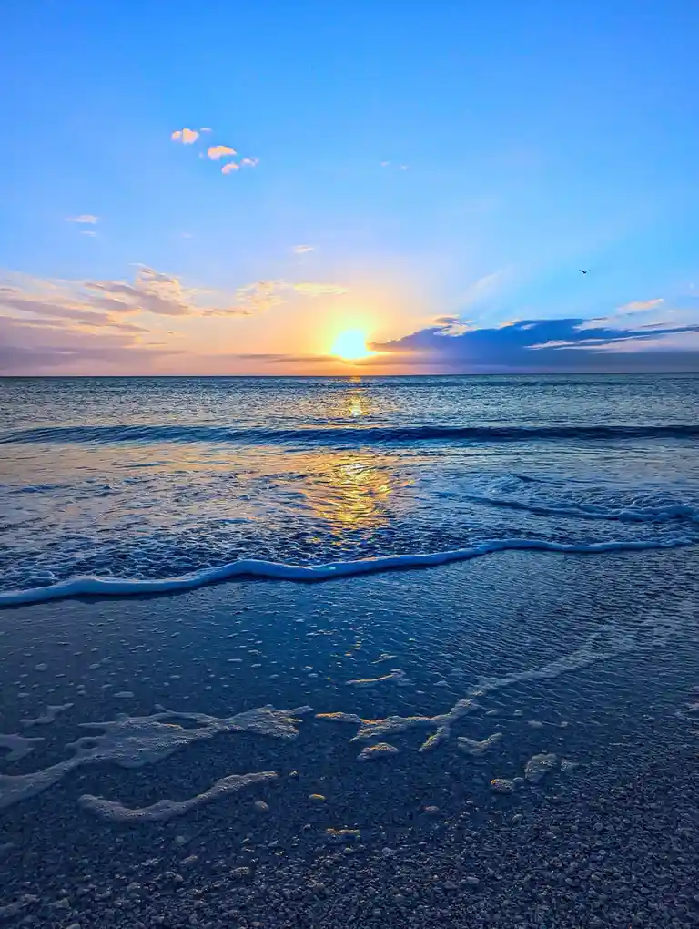 A serene beach sunset photo with the sun low on the horizon, casting golden light across the water and wet sand. Gentle…