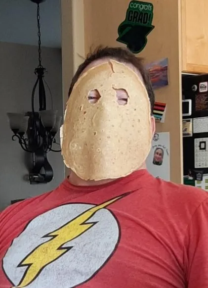 A person wearing a red 'The Flash' t-shirt has a tortilla mask covering their face, with eye holes cut out. The background…