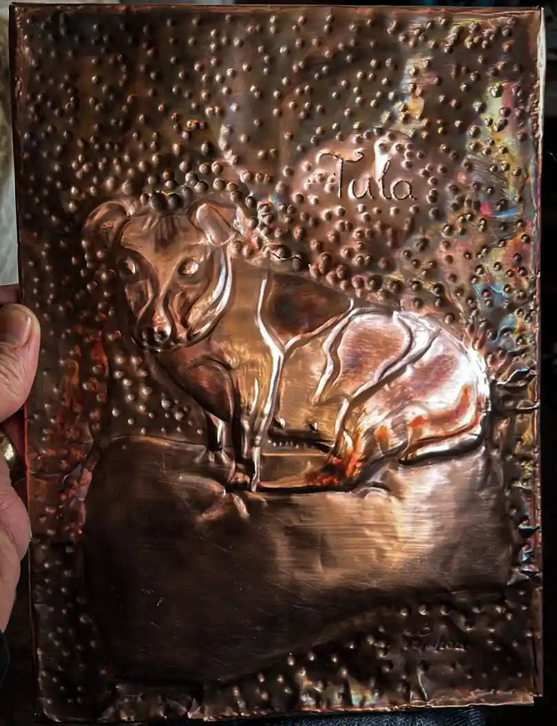 A hand holds a hammered copper plaque featuring a raised, stylized relief of a dog, with the name 'Tula' engraved above it…