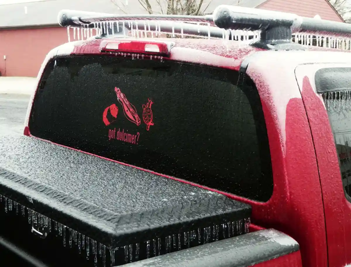 The rear of a red pickup truck is covered in snow and ice, with icicles hanging from the roof rack and tailgate. A red decal…