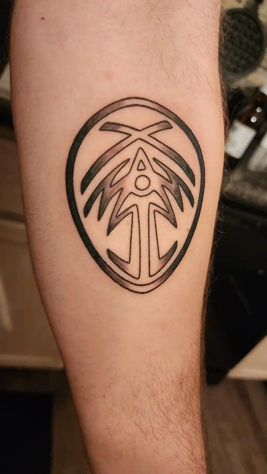 A close-up photo shows a black-and-gray tattoo on a person's forearm, depicting a stylized, symmetrical tribal or geometric…