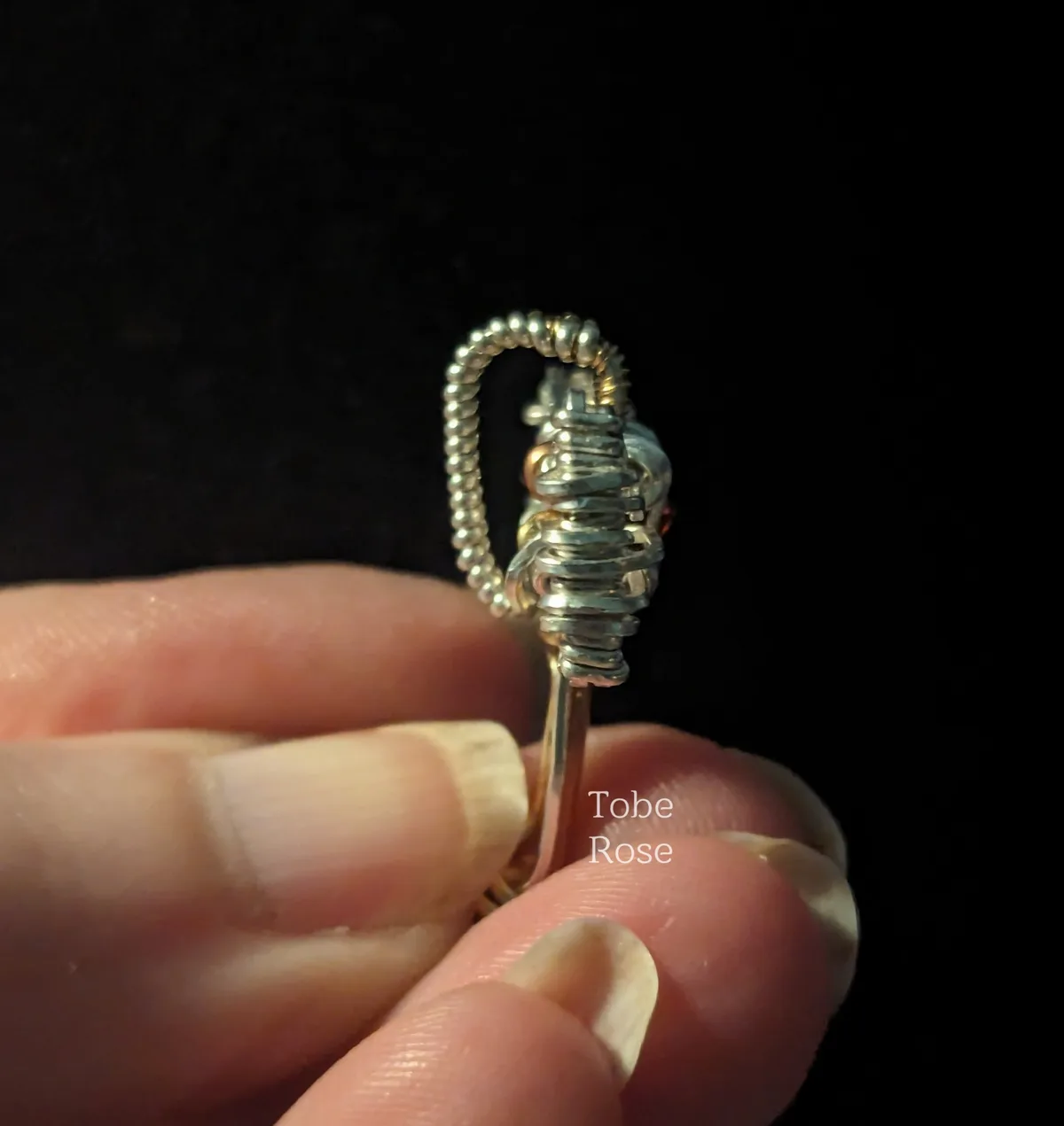 A close-up photo shows a hand holding a small, intricate piece of wire-wrapped jewelry against a black background. The…