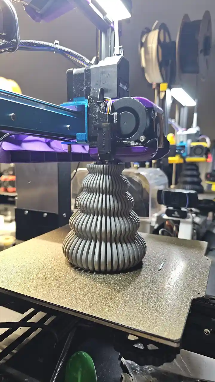 A 3d printer with a purple and black extruder is actively printing a gray, vertically ribbed, hourglass-shaped object on a…