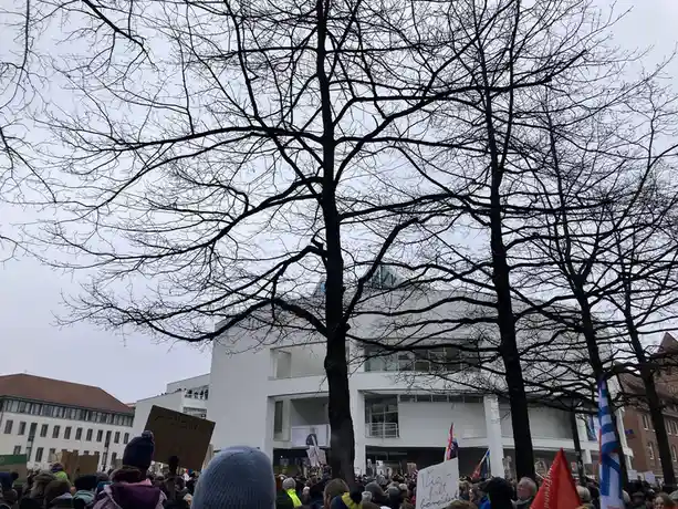 A crowd of protesters holding signs and banners stands in front of a modern white building, framed by the bare, intricate…