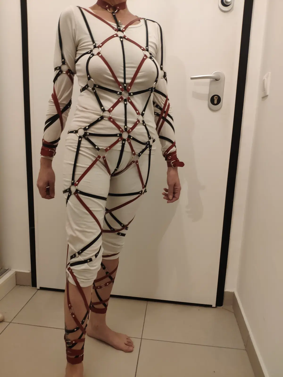 A person stands in front of a white door wearing a full-body white outfit overlaid with an intricate web of red and black…