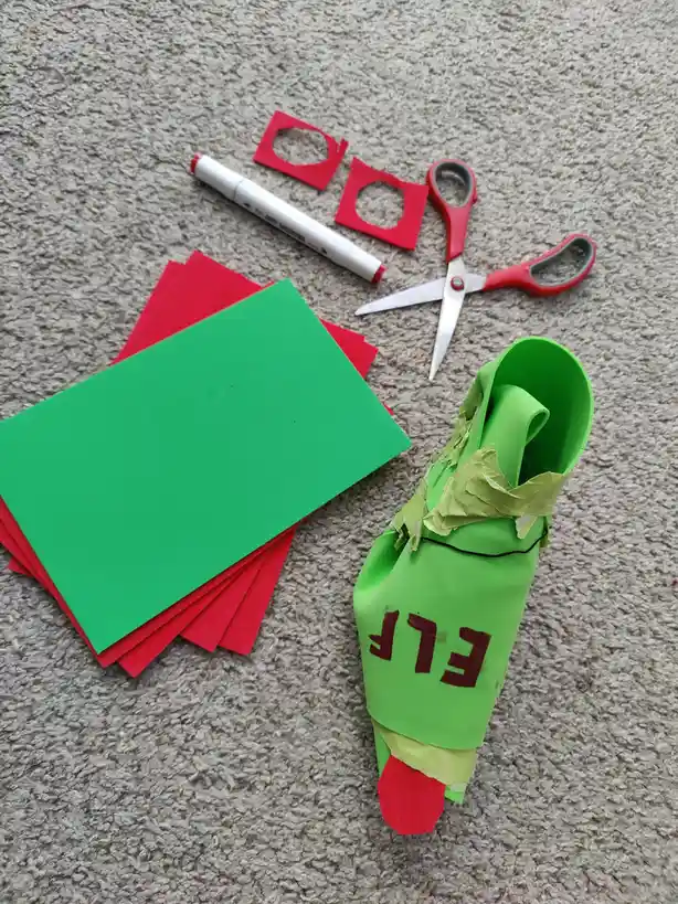 A handmade green paper elf shoe with red felt toe and 'ELF' written in red marker lies on carpet next to crafting supplies…