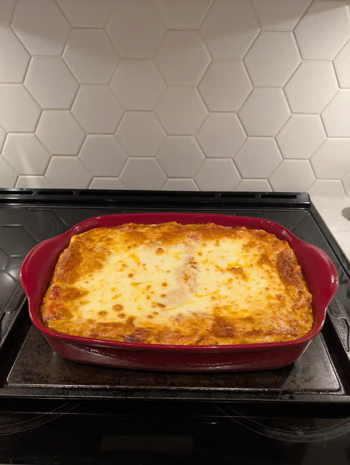 A red rectangular baking dish filled with golden-brown, bubbly cheese-topped lasagna sits on a black electric stovetop, with…