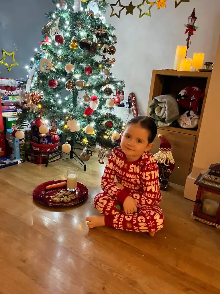 A young child in red holiday pajamas sits cross-legged on a wooden floor in front of a decorated Christmas tree, looking at…