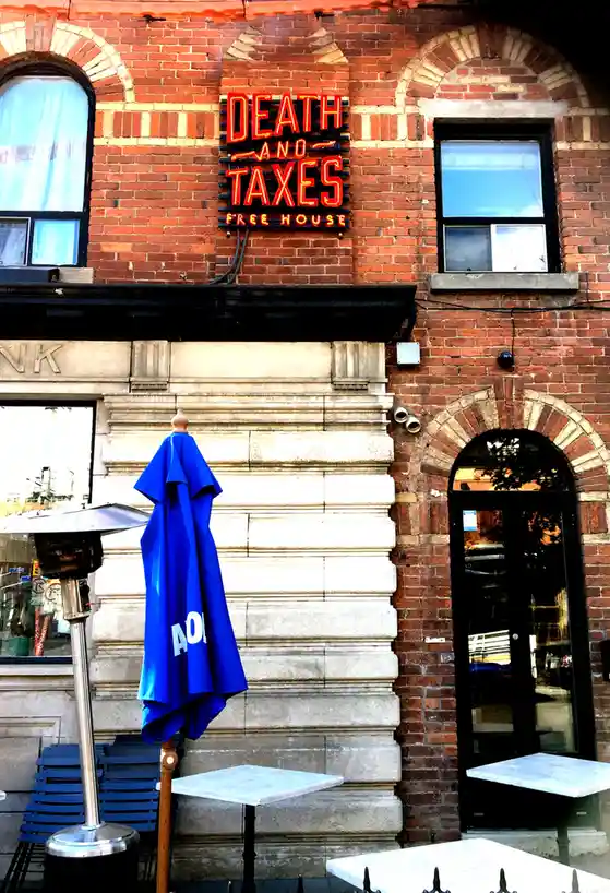 A red brick building with arched windows and a black door, featuring a bright red neon sign that reads 'DEATH AND TAXES FREE…