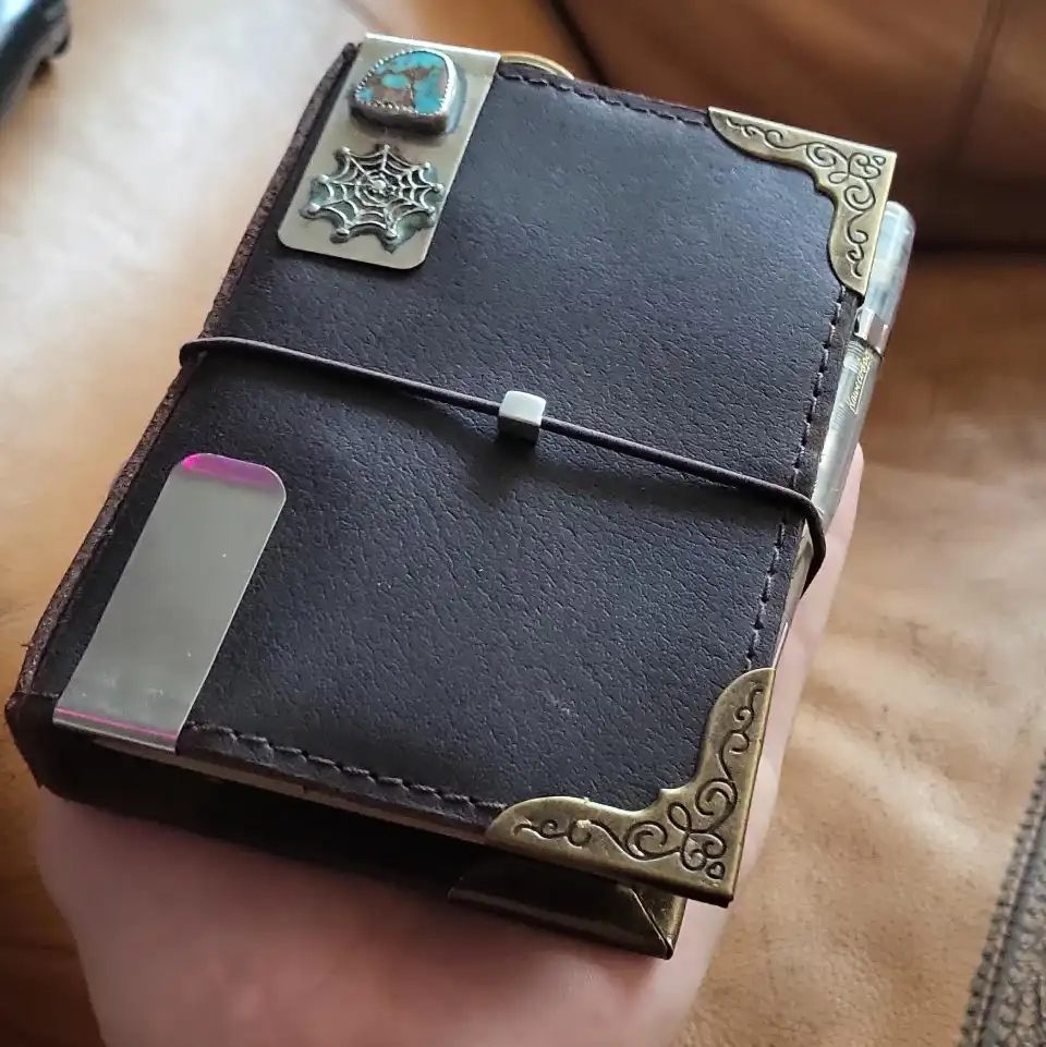 A hand holds a small, dark brown leather-bound journal with ornate brass corner guards and a decorative metal plate…