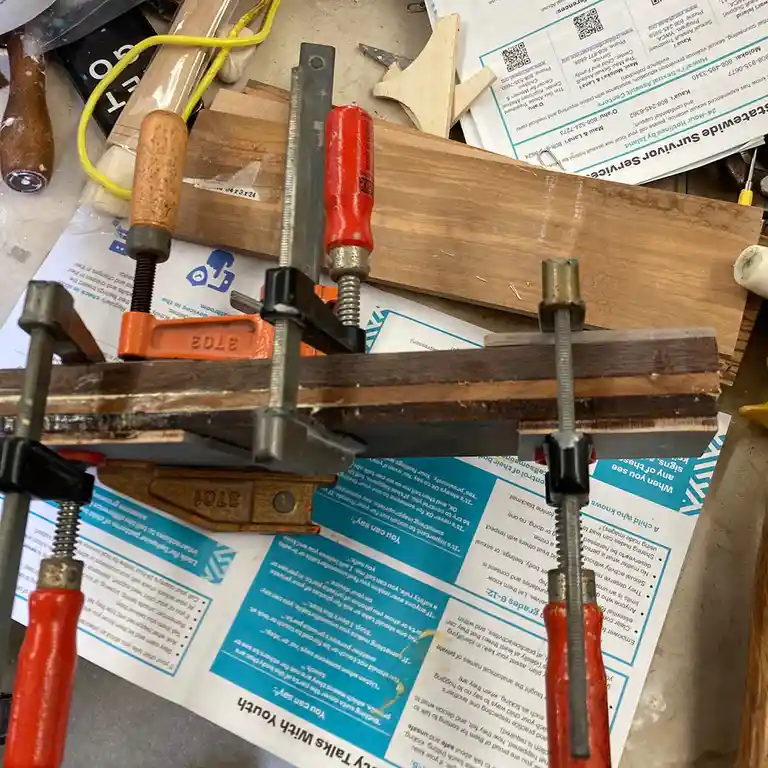 A cluttered workbench shows several woodworking clamps holding pieces of wood together, with multiple printed brochures…