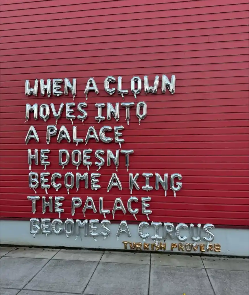 Silver foil balloons spell out a humorous proverb on a red corrugated metal shutter, stating that when a clown moves into a…
