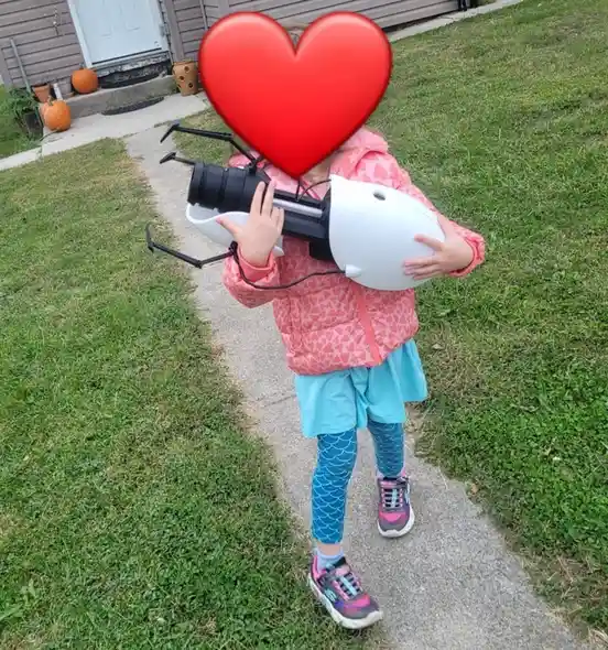 A young child wearing a pink heart-patterned jacket and blue leggings stands on a sidewalk in a grassy yard, holding a large…