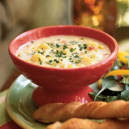 a red pedestal bowl filled with creamy corn chowder, garnished with chopped green herbs, sits on a green plate with…