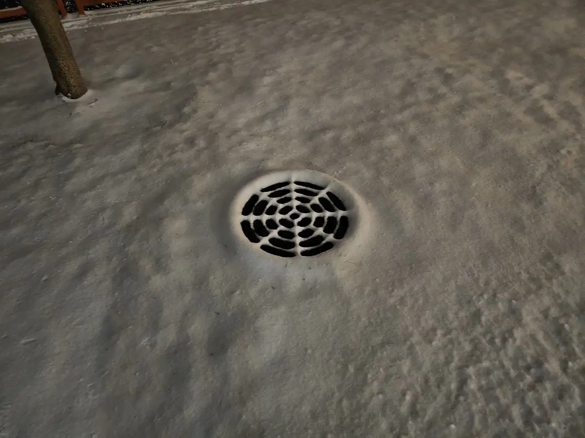 A close-up photo shows a circular metal storm drain grate set into a snowy, icy sidewalk. The grate's pattern of oval holes…