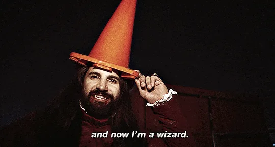 A man with a long beard and dark clothing wears an orange traffic cone on his head, holding it with one hand while his…