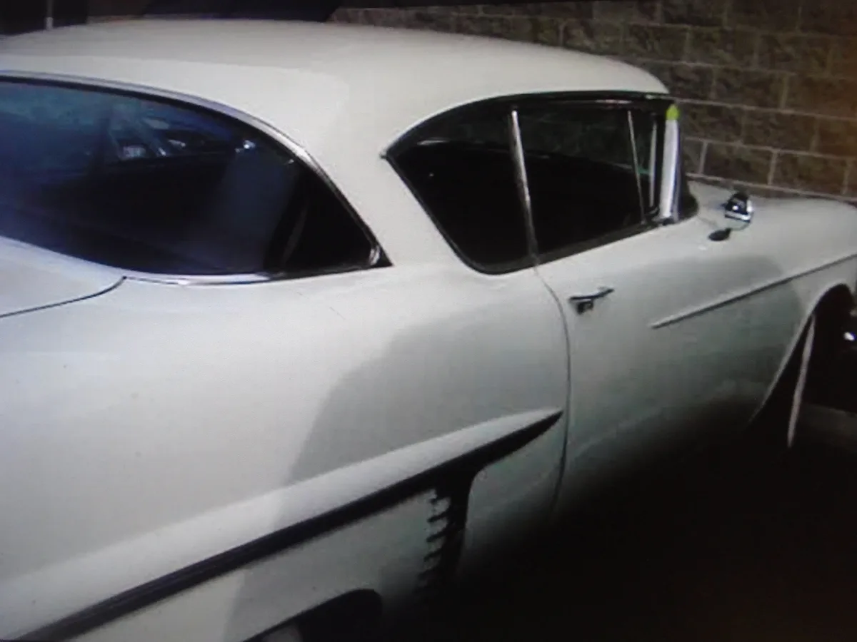 A low-resolution photo shows the rear passenger side of a white classic American car, likely from the 1950s or 60s, parked…