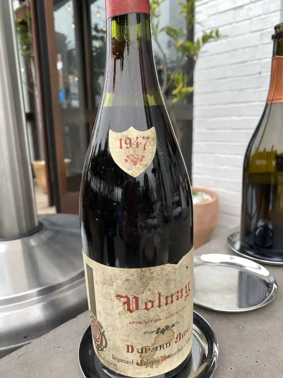 A close-up photo of an old, dusty wine bottle labeled 'Volnay' with a shield-shaped sticker reading '1947' on its neck…